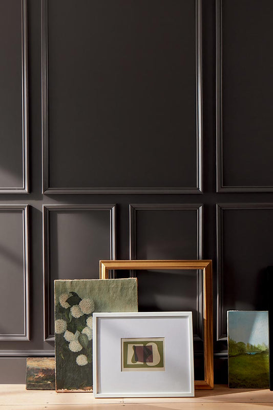 Artwork and frames arranged on a floor against a deep chocolate brown-painted wall with decorative molding.